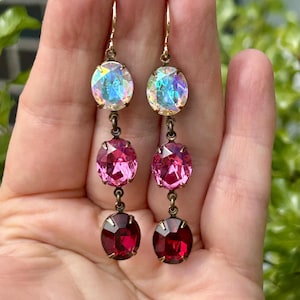 May include: Dangle earrings featuring three oval gemstones in iridescent, pink, and deep red hues. The earrings are suspended from gold-coloured hooks. Each stone is approximately 1.5 cm in length, creating a striking contrast.