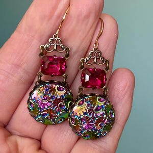 May include: Vintage-style dangle earrings with a gold-tone metal setting. Each earring features a rectangular, vibrant pink gemstone above a round, iridescent, floral-patterned charm. The earrings have a decorative filigree design and a hook closure.