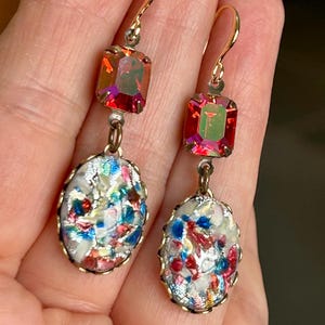 Peut inclure: Une paire de boucles d'oreilles pendantes avec une pierre précieuse rouge irisée et un charme ovale blanc avec un motif de confettis colorés.