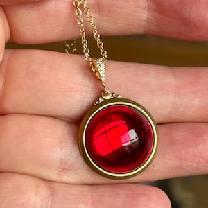 May include: A gold chain necklace with a pendant featuring a large, round, red glass cabochon set in a gold frame.