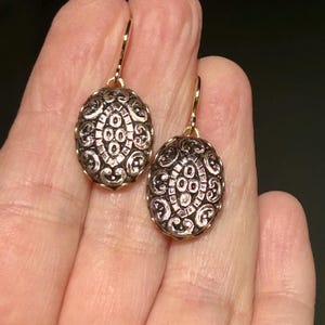Può includere: Un paio di orecchini ovali con intricati disegni in filigrana d'argento e ganci dorati. Gli orecchini presentano un motivo dettagliato e ornato, con un disegno centrale di cerchi e linee. Lo sfondo è scuro.