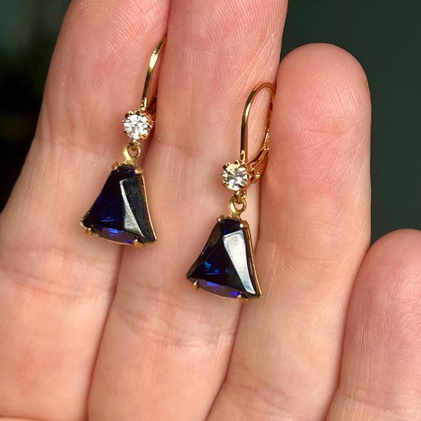 Könnte beinhalten: Ein Paar goldfarbene Ohrringe mit einem dreieckigen, tiefblauen Edelsteinanhänger. Jeder Ohrring hat einen kleinen, runden, klaren Stein über dem Anhänger. Die Ohrringe werden mit einem goldfarbenen Bügelverschluss gehalten.
