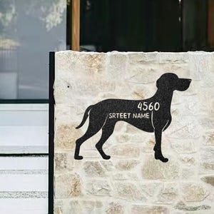May include: Black metal house number sign in the shape of a dog. The sign displays the number "4560" and the street name. The sign is mounted on a textured, light-colored stone wall.