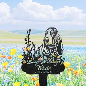 May include: Black metal garden stake with a dog silhouette, wildflowers, and the inscription "In Loving Memory Of Trixie 2012-2024." The dog's head is detailed with etched lines. The stake is set in a field of colorful flowers under a blue sky.