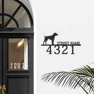 Puede incluir: Letrero de dirección de metal negro con una silueta de perro, las palabras "Street Name" y los números "4321". El letrero está montado en una pared blanca. El letrero está cerca de una puerta y algo de vegetación.