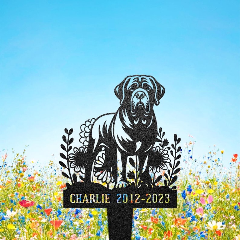 May include: Black metal dog memorial stake with a Mastiff silhouette, floral and leaf cutouts. The stake has the name "CHARLIE" and the years "2012-2023". The background is a colourful wildflower meadow and a blue sky.