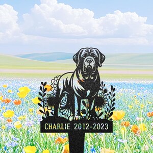 May include: Black metal dog memorial stake with a Mastiff silhouette and floral details. The stake is inscribed with "Charlie 2012-2023". The background depicts a field of wildflowers under a blue sky with clouds.