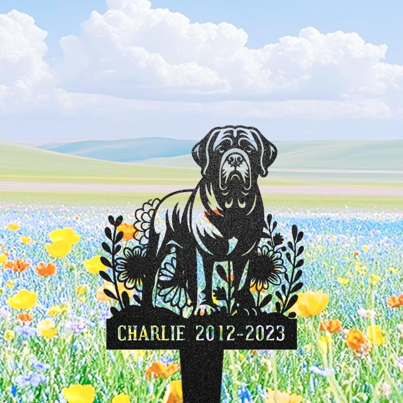 May include: Black metal dog memorial stake with a Mastiff silhouette and floral details. The stake is inscribed with "Charlie 2012-2023". The background depicts a field of wildflowers under a blue sky with clouds.