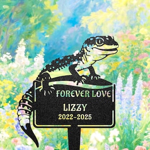 May include: A black metal garden stake featuring a gecko design. The stake has the words "FOREVER LOVE" and the name "LIZZY" with the years "2022-2025". The gecko is black with blue and yellow accents, set against a colorful floral background.