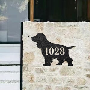 以下が含まれることがあります： 犬の形をした黒い金属製のサインで、中央に1028の数字があります。明るい色の石壁に取り付けられています。犬のシルエットはテクスチャ加工されています。