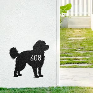 May include: Black metal silhouette of a dog with the number 608 in white. The dog is standing on a white wall, with a grassy area in the foreground. The dog's tail is curled, and the number is centered on its body.