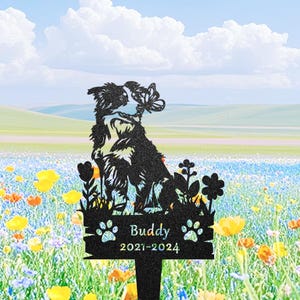 Puede incluir: Estaca de jardín de metal negro con la silueta de un perro con una mariposa. La estaca incluye el nombre "Buddy" y los años "2021-2024". El fondo muestra un campo de flores silvestres bajo un cielo azul.