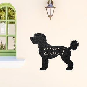 Puede incluir: Silueta metálica negra de un perro, posiblemente un caniche, con el año "2007" recortado en el cuerpo. El perro está colocado sobre una pared color crema. Una ventana verde y una lámpara de pared son visibles en el fondo.