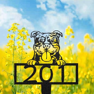 Könnte beinhalten: Ein schwarzer Metall-Gartenstecker mit einem lächelnden Bulldoggen, die Zunge herausstreckt und ein Schild mit der Nummer 201 hält. Der Hintergrund ist ein Feld mit gelben Blumen unter blauem Himmel.