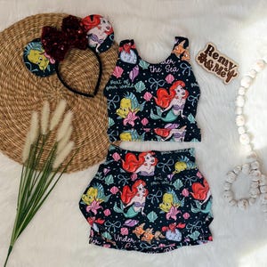 Puede incluir: Un conjunto negro de dos piezas con un estampado de sirena con Ariel, Flounder y Sebastián. El conjunto incluye un top corto y una falda. También hay una diadema a juego con orejas de lentejuelas y un letrero de madera que dice "Remy & Mey".