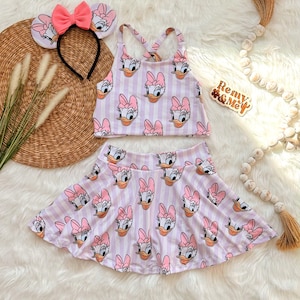 Puede incluir: Un conjunto infantil con estampado de Daisy Duck. El conjunto incluye un top corto y una falda skater con una diadema a juego. El conjunto es de rayas blancas y lavanda con la cara de Daisy Duck y un lazo rosa. La diadema tiene un lazo rosa y orejas de Daisy Duck.