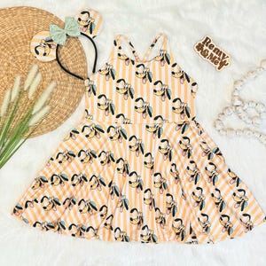 May include: A yellow and white striped dress featuring a Pluto character print. The dress has a flared skirt and crisscross straps. A matching headband with Pluto ears and a mint green bow is also present. A wooden sign that says "Remy & Me" is also present.