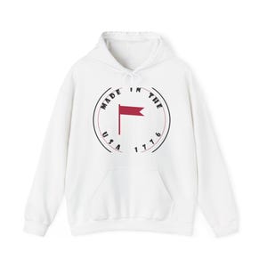 May include: A white long-sleeve hooded sweatshirt featuring a central circular graphic. The design includes the text "MADE IN THE USA 1776" in black lettering arranged around a red flag icon centred within the circle on the front of the garment.