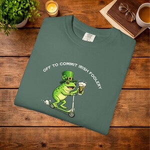 May include: Green t-shirt with the phrase "OFF TO COMMIT IRISH FOOLERY" and a cartoon frog wearing a leprechaun hat, riding a scooter, and holding a beer. The shirt is made of a soft material.