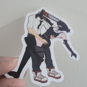 May include: A sticker featuring a stylized illustration of two figures in a dynamic pose. One figure has a chainsaw head and is holding the other figure, who is wearing a black dress and boots. The sticker has a white border.