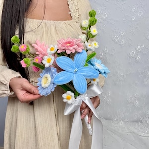 May include: A bouquet of handmade fabric flowers in shades of pink, blue, and white. The bouquet features large blue flowers, pink daisies, and smaller white blossoms, tied with a white satin ribbon. The flowers are held by a person wearing a beige dress.