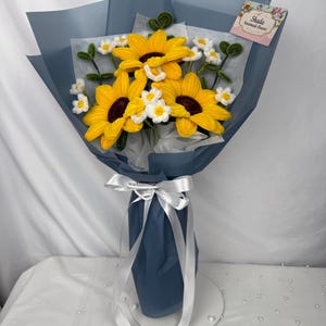 May include: A bouquet of handmade crochet flowers, featuring bright yellow sunflowers and small white daisies. The bouquet is wrapped in blue paper and tied with a white ribbon. A tag reads "Shada Handmade Flowers".