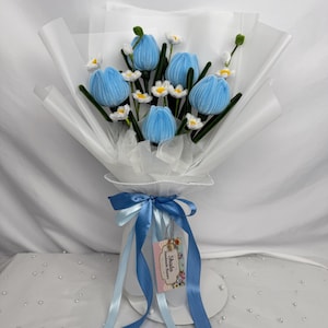 Op de afbeelding: Een boeket handgemaakte blauwe tulpen met witte madeliefjes accenten, verpakt in wit papier. Het boeket is vastgebonden met een blauw lint en een label met de tekst "Shada Handmade Flowers".