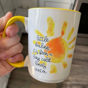 May include: White ceramic mug with a yellow rim and handle. The mug features a yellow and orange sun design with the text "little darling, it's been a long cold lonely winter".