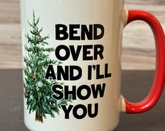 May include: White ceramic coffee mug with a red rim and handle. The mug has a Christmas tree design and the text "Bend over and I'll show you".