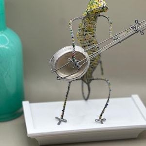 May include: A whimsical sculpture of a lizard playing a banjo. The lizard is crafted from wire and adorned with colorful beads. The banjo is made from a bottle cap. The sculpture is on a white platform.