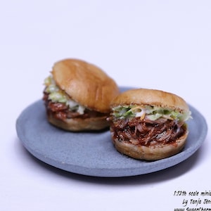 Könnte beinhalten: Zwei Miniatur-Pulled-Pork-Sandwiches mit Krautsalat auf einem grauen Teller. Die Sandwiches haben goldbraune Brötchen und sind eine Miniatur im Maßstab 1:12. Das Bild enthält den Text "1:12th scale miniatures by Tanja Jensen".