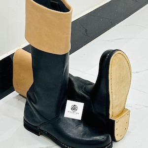 May include: A pair of black leather boots featuring a tall, tan-colored cuff at the top. The boots have a flat, light-colored sole with visible stitching along the edge. A small white square tag with the text ATHLETIGO is attached to the side.