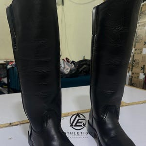 Puede incluir: Un par de botas altas de cuero negro con diseño de punta cuadrada y tacón bajo. Las botas presentan un acabado liso, una caña estructurada que llega por debajo de la rodilla y costuras visibles a lo largo de la suela. Se observa un logotipo con el texto ATHLETIGO ENTERPRISES.
