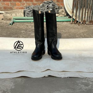 May include: A pair of black knee-high boots with a smooth finish, rounded toes, and a low heel. The boots feature a tall, structured shaft and visible stitching along the sole. A logo with the text ATHLETIGO ENTERPRISES is visible to the left.