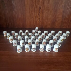 May include: A collection of white porcelain thimbles, each adorned with a portrait of a woman. The thimbles are arranged on a brown surface, showcasing a variety of historical figures and elegant designs. The thimbles are a collectible item.