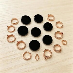 May include: A collection of jewelry components consisting of six smooth, circular black discs and several rose gold-toned metal rings, some featuring small loops or clover-shaped connectors, arranged in a scattered pattern on a light surface.