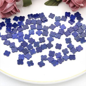 May include: A collection of small, four-petal flower-shaped lapis lazuli beads. The beads are a deep blue color and are scattered on a white surface. The image is well-lit, highlighting the texture and color of the beads.
