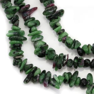 May include: Close-up of a necklace made of green and red-toned gemstone beads. The beads are irregularly shaped and strung together, creating a natural and earthy aesthetic. The necklace is displayed against a white background.