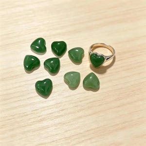 May include: A collection of eight small, smooth, heart-shaped green stones alongside a single metal ring featuring a matching green heart-shaped stone set into the band. The stones vary slightly in their shade of green, ranging from deep forest to lighter sage tones.
