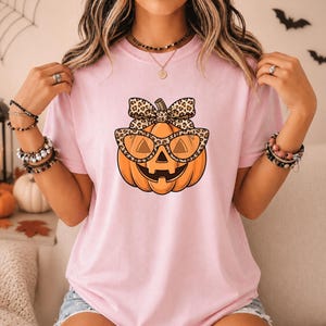 May include: A light pink short-sleeve t-shirt featuring a central graphic of an orange pumpkin wearing leopard print glasses and a matching leopard print bow on top. The pumpkin has a carved face with triangular eyes and a wide, toothless grin.