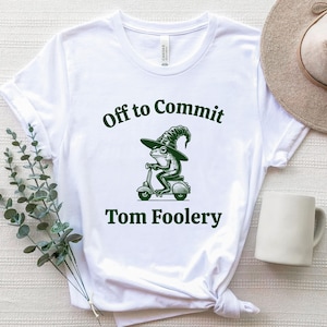 Könnte beinhalten: Weißes T-Shirt mit einem grünen Aufdruck einer Frosch-Illustration mit Hexenhut auf einem Roller. Der Text "Off to Commit Tom Foolery" steht darüber und darunter.
