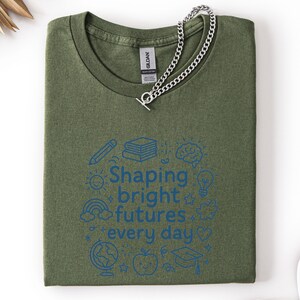 May include: Olive green t-shirt with the text "Shaping bright futures every day" in blue, surrounded by school-themed illustrations. A silver chain necklace with a toggle clasp rests on the shirt.