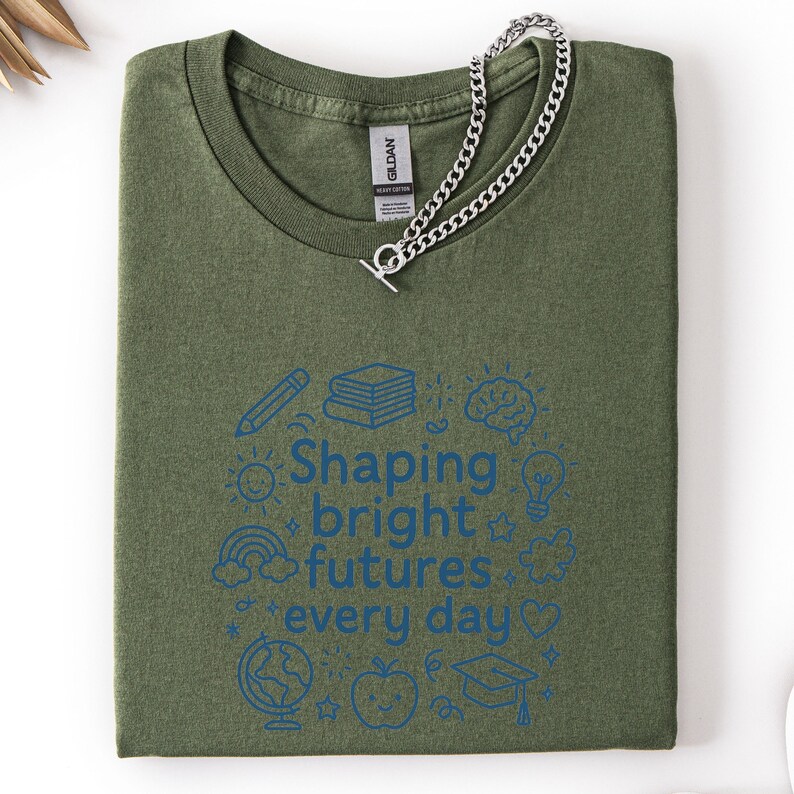 May include: Olive green t-shirt with the text "Shaping bright futures every day" in blue, surrounded by school-themed illustrations. A silver chain necklace with a toggle clasp rests on the shirt.