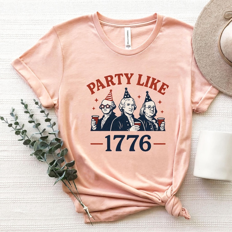 K&ouml;nnte beinhalten: Hellrosa T-Shirt mit dem Schriftzug "PARTY LIKE" in Rot &uuml;ber einer Illustration von drei M&auml;nnern in Kolonialkleidung, die jeweils eine Tasse halten. Die Zahl "-1776-" ist darunter in Marineblau gedruckt.