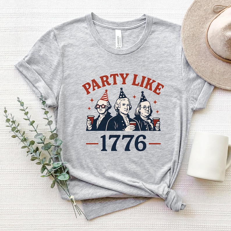 K&ouml;nnte beinhalten: Ein meliertes graues T-Shirt mit dem Text "PARTY LIKE 1776" in Rot und Marineblau. Die Grafik zeigt drei historische Figuren mit Partyh&uuml;ten, die rote Becher halten. Ein l&auml;ssiges Kleidungsst&uuml;ck.