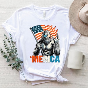 May include: White t-shirt featuring a graphic of a Bigfoot-like creature wearing sunglasses and holding an American flag. The word "'MERICA" is printed in red, white, and blue below the image. The creature is giving a hand gesture.