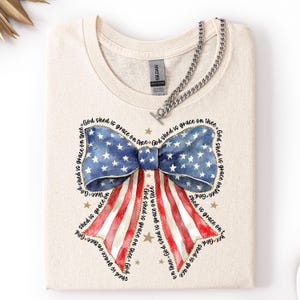 May include: A folded, cream-colored t-shirt featuring a patriotic bow design. The bow is a watercolor illustration of the American flag, with blue stars and red and white stripes. The text "God shed is grace on thee" surrounds the bow.