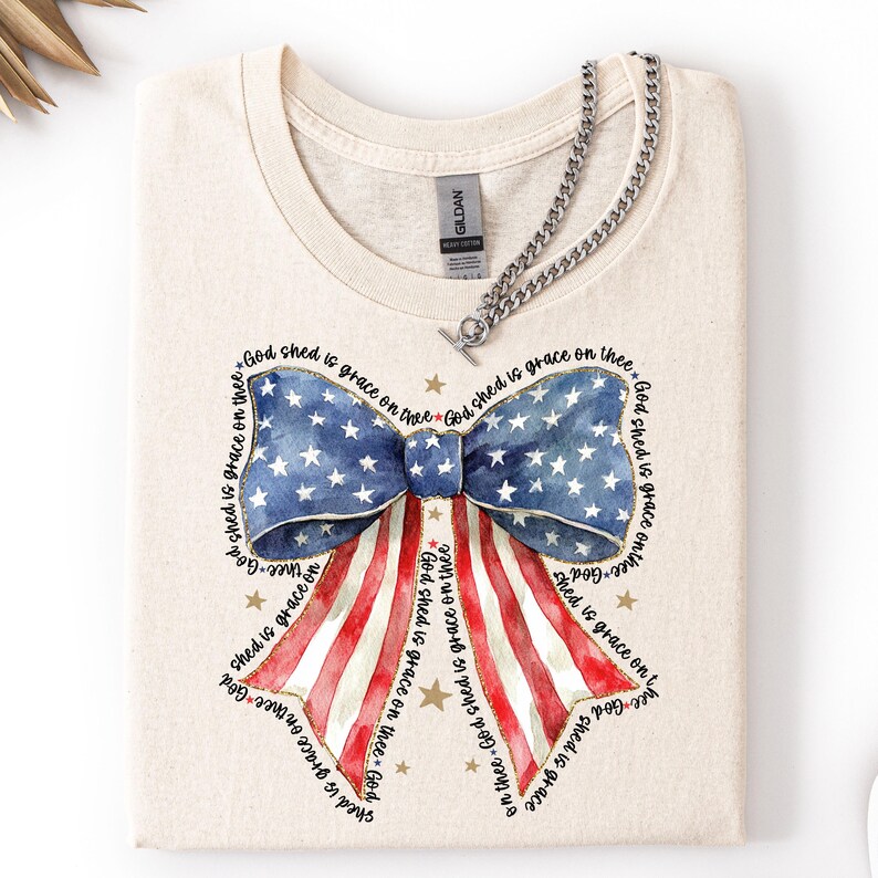 May include: A folded, cream-colored t-shirt featuring a patriotic bow design. The bow is a watercolor illustration of the American flag, with blue stars and red and white stripes. The text "God shed is grace on thee" surrounds the bow.