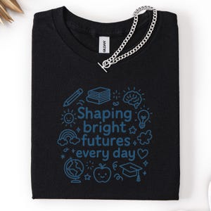 May include: Black t-shirt with a blue graphic that reads "Shaping bright futures every day" surrounded by illustrations of a pencil, books, brain, lightbulb, rainbow, globe, apple, and graduation cap. A silver chain is draped across the shirt.