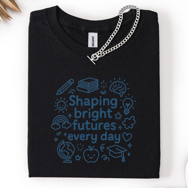 May include: Black t-shirt with a blue graphic that reads "Shaping bright futures every day" surrounded by illustrations of a pencil, books, brain, lightbulb, rainbow, globe, apple, and graduation cap. A silver chain is draped across the shirt.